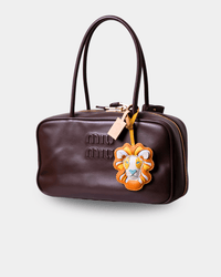 A dark brown leather Miu Miu handbag with a top handle, gold-tone zipper, and a lion patch charm, shown on a light gray background.