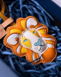 A close-up of a handmade leather accessory, likely a belt buckle or decorative plaque, featuring a stylized lion face. The piece is crafted from orange, yellow, and off-white leather with light blue embroidered details, resting on blue paper shred inside a dark box.