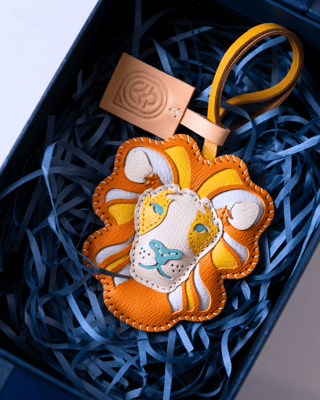 A handmade lion-shaped leather keychain or bag charm in orange, yellow, and white, displayed inside a dark box filled with blue paper shred. A tan leather tag with an embossed logo is attached.