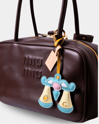 A close-up of a dark brown Miu Miu leather handbag featuring an embroidered celestial charm in light blue, cream, pink, and yellow, with gold-tone hardware and a beige brand tag.