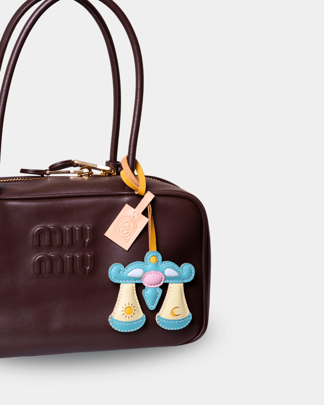 A close-up of a dark brown leather Miu Miu handbag featuring a decorative embroidered alien charm with a peach tag.
