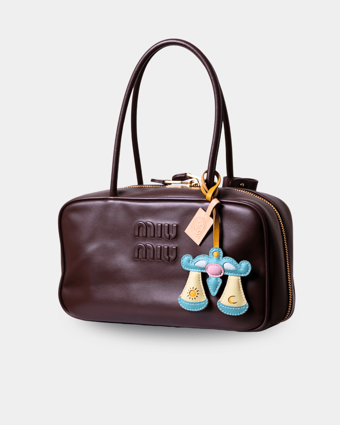 A dark brown Miu Miu leather handbag with gold-tone hardware, featuring the embossed Miu Miu logo on the front, dual top handles, and a decorative blue and yellow boot-shaped charm with a pink flower accent.