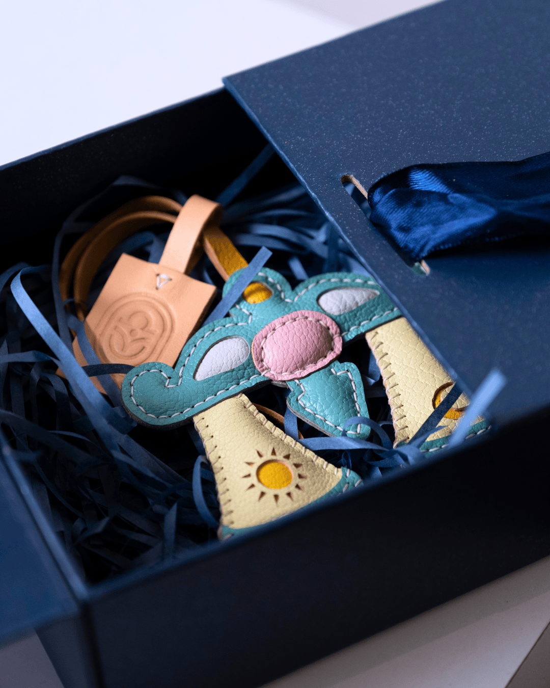 A handmade leather koala ornament inside a dark blue gift box with shredded paper filler and a leather tag.