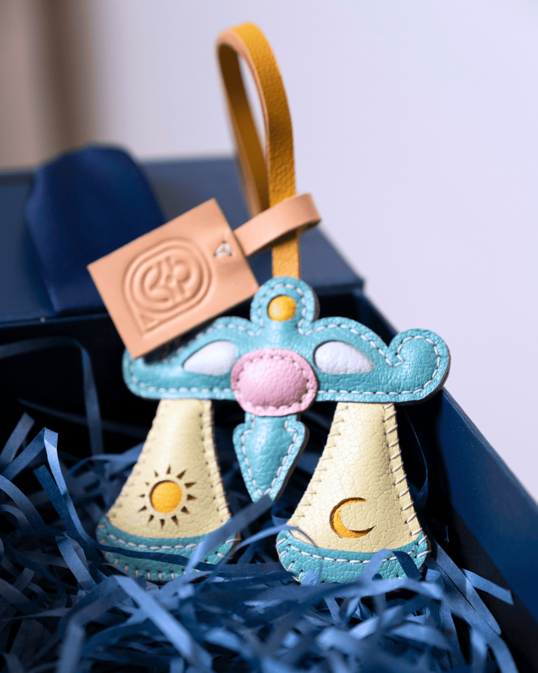 A handmade leather celestial charm set featuring a crescent moon and sun design in light blue and cream, nestled in blue paper shred inside a dark gift box.