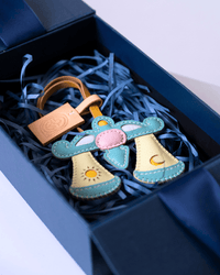 A handmade leather angel keychain with a light brown leather strap and a matching embossed leather tag, displayed inside a dark blue gift box filled with blue paper shred. The angel features light blue wings, a pink halo, and cream-colored halos with embroidered sun and moon details.