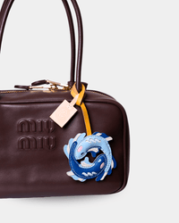 A close-up of a dark brown leather Miu Miu handbag featuring a signature blue and white dragon charm attached with a yellow ribbon and a small beige paper tag.