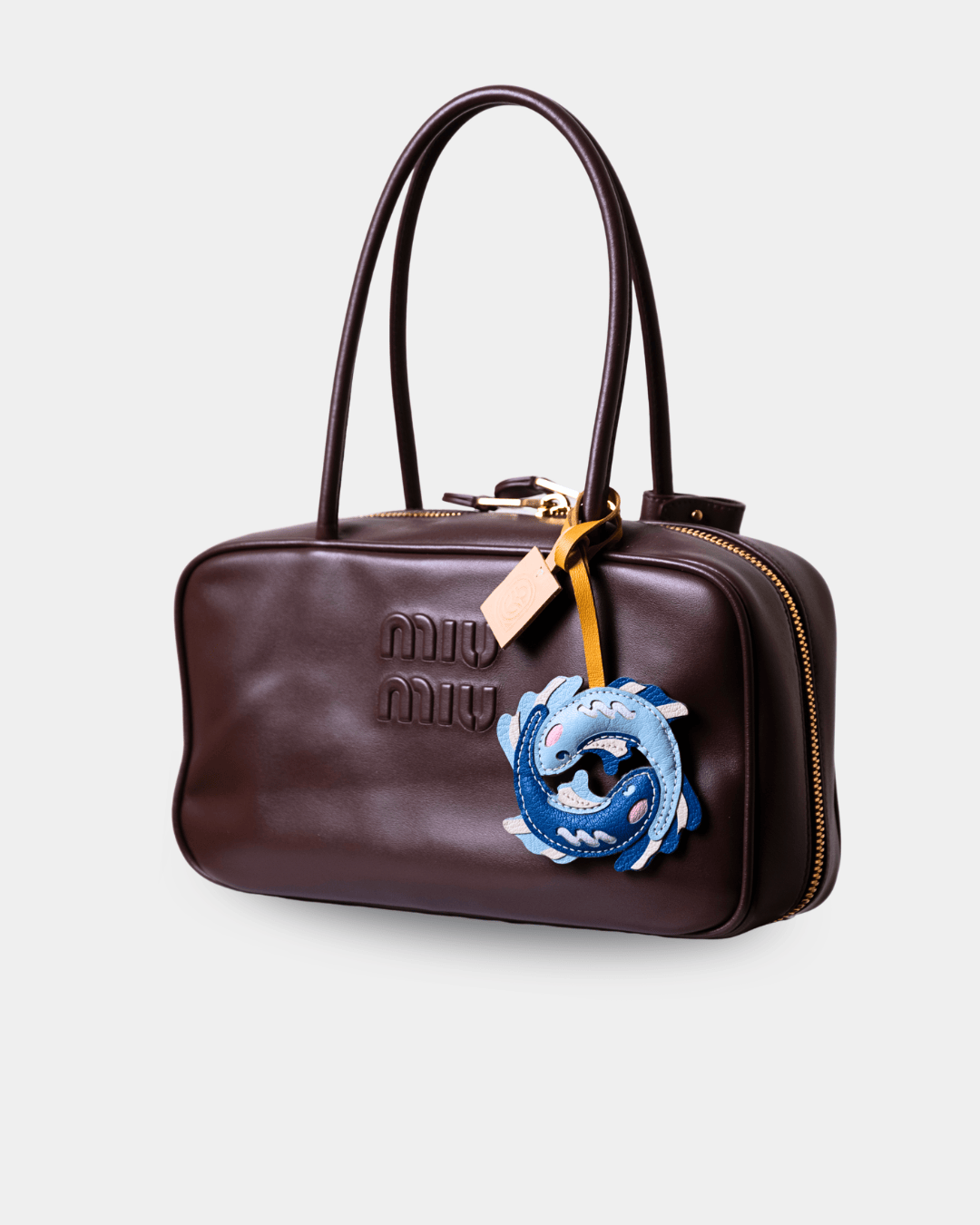 A dark brown leather Miu Miu handbag with a top handle, gold-tone zipper, and a blue dragon charm attached to a beige leather tag, shown against a light gray background.
