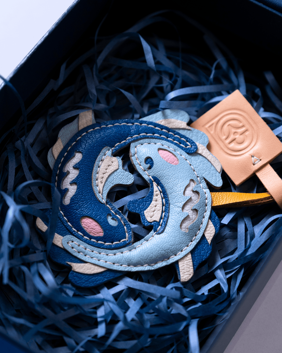 A close-up of a leather keychain or charm shaped like two interlocking dolphins in shades of blue, with beige stitching and pink accents, resting in dark blue shredded paper inside a gift box next to a tan leather tag.