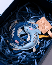 A close-up of a leather keychain or charm shaped like two interlocking dolphins in shades of blue, with beige stitching and pink accents, resting in dark blue shredded paper inside a gift box next to a tan leather tag.