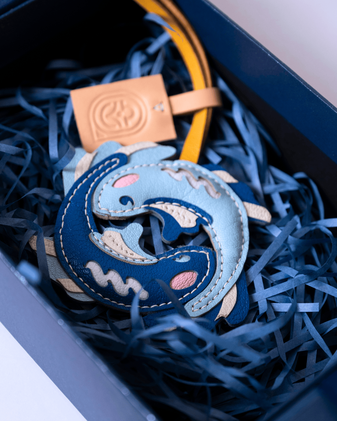 A close-up of a blue and navy leather keychain or bag charm with a stylized animal design, placed in blue shredded paper inside a dark box. A tan leather tag with an embossed logo is attached, along with a yellow strap.