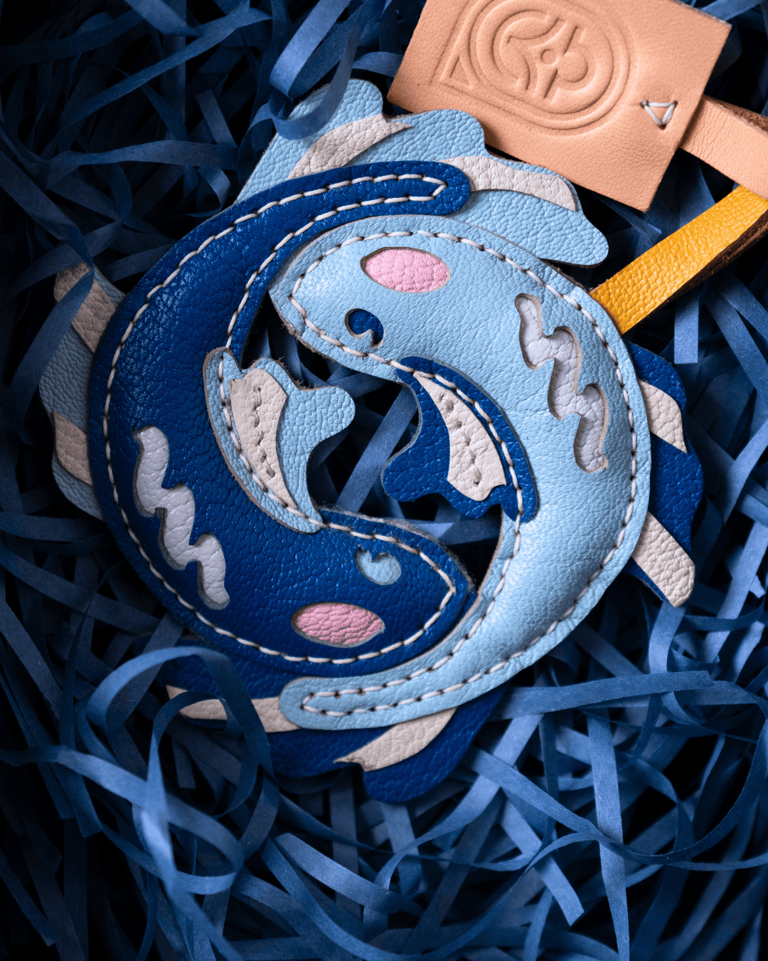 A close-up of a leather keychain charm shaped like two interlocking dolphins in shades of blue, cream, and pink, resting on blue shredded paper with a tan leather tag partially visible.