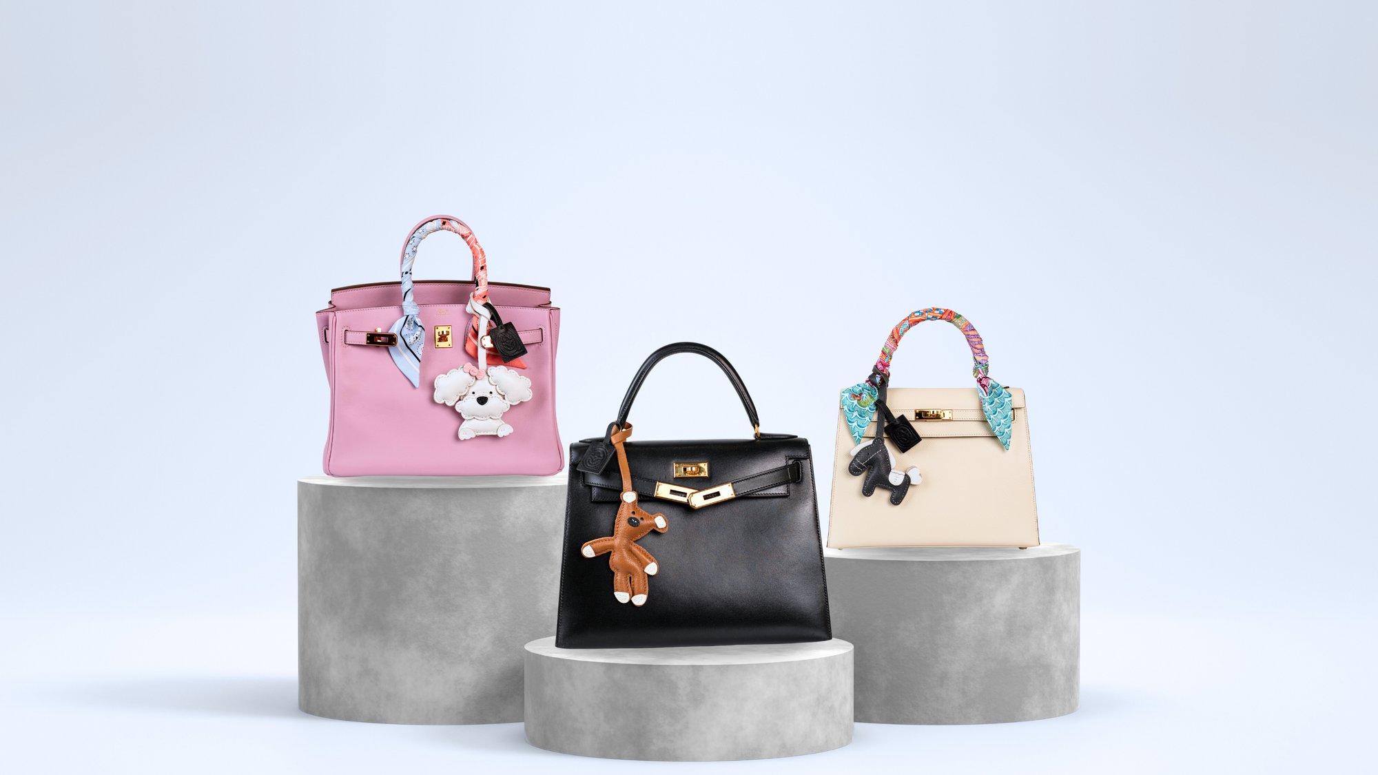 Three designer handbags with luxury leather charms on concrete stands against a light blue background
