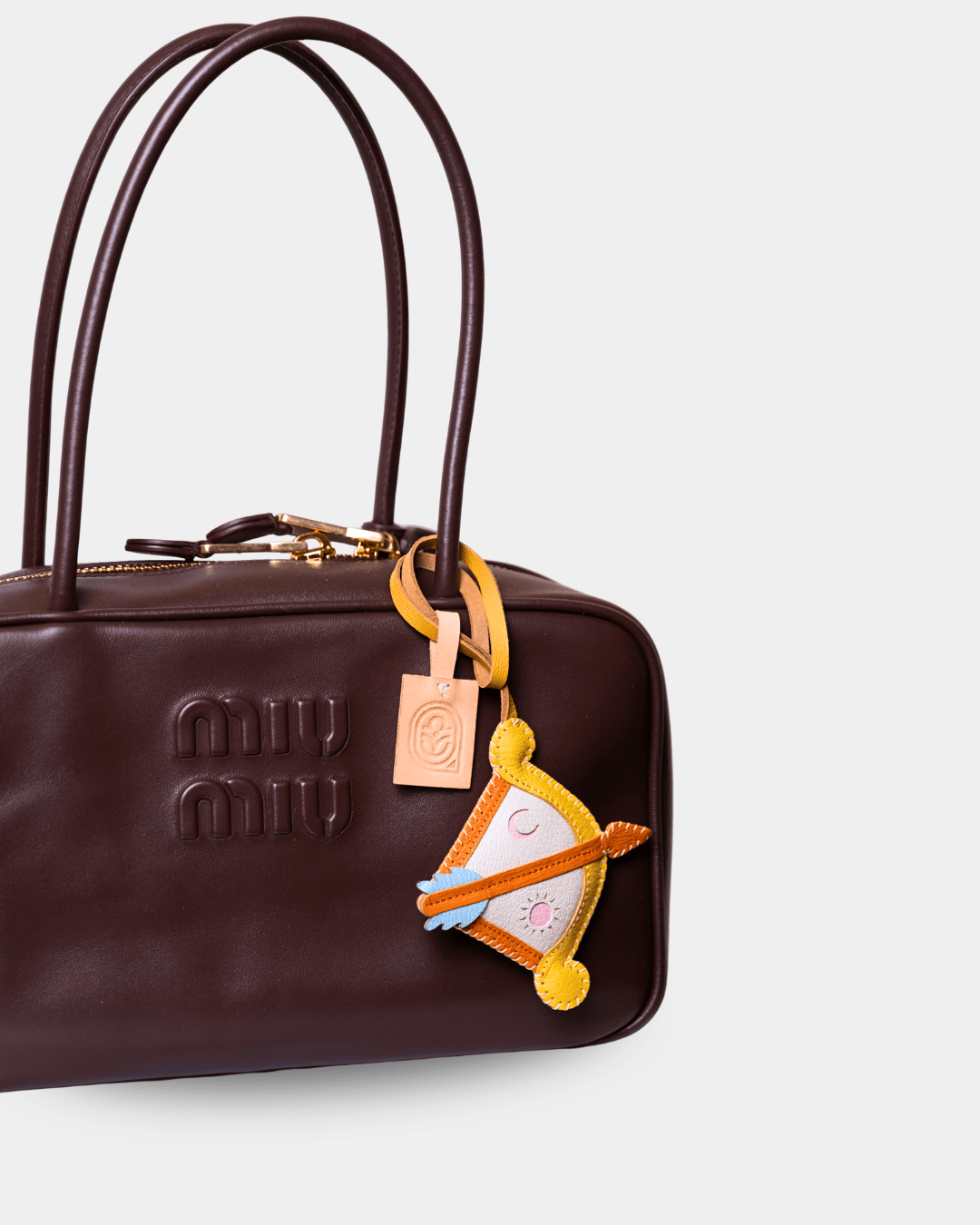 A dark brown leather Miu Miu handbag with gold-tone hardware, featuring the embossed Miu Miu logo on the front and a decorative yellow and orange horse-shaped charm.