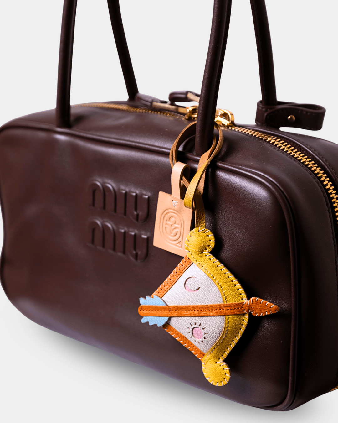 A close-up of a dark brown leather Miu Miu handbag with gold-tone hardware, featuring an embroidered bow and arrow keychain charm.
