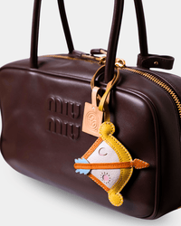 A close-up of a dark brown leather Miu Miu handbag with gold-tone hardware, featuring an embroidered bow and arrow keychain charm.
