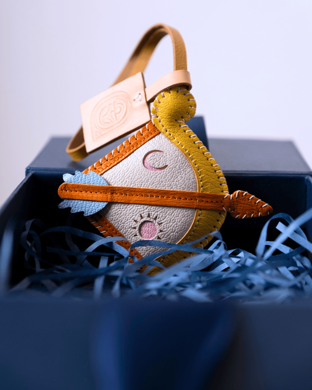 A handmade leather crescent moon ornament with a bird design, featuring yellow and white leather, orange stitching, and a tan strap, nestled in blue paper shred inside a dark box.
