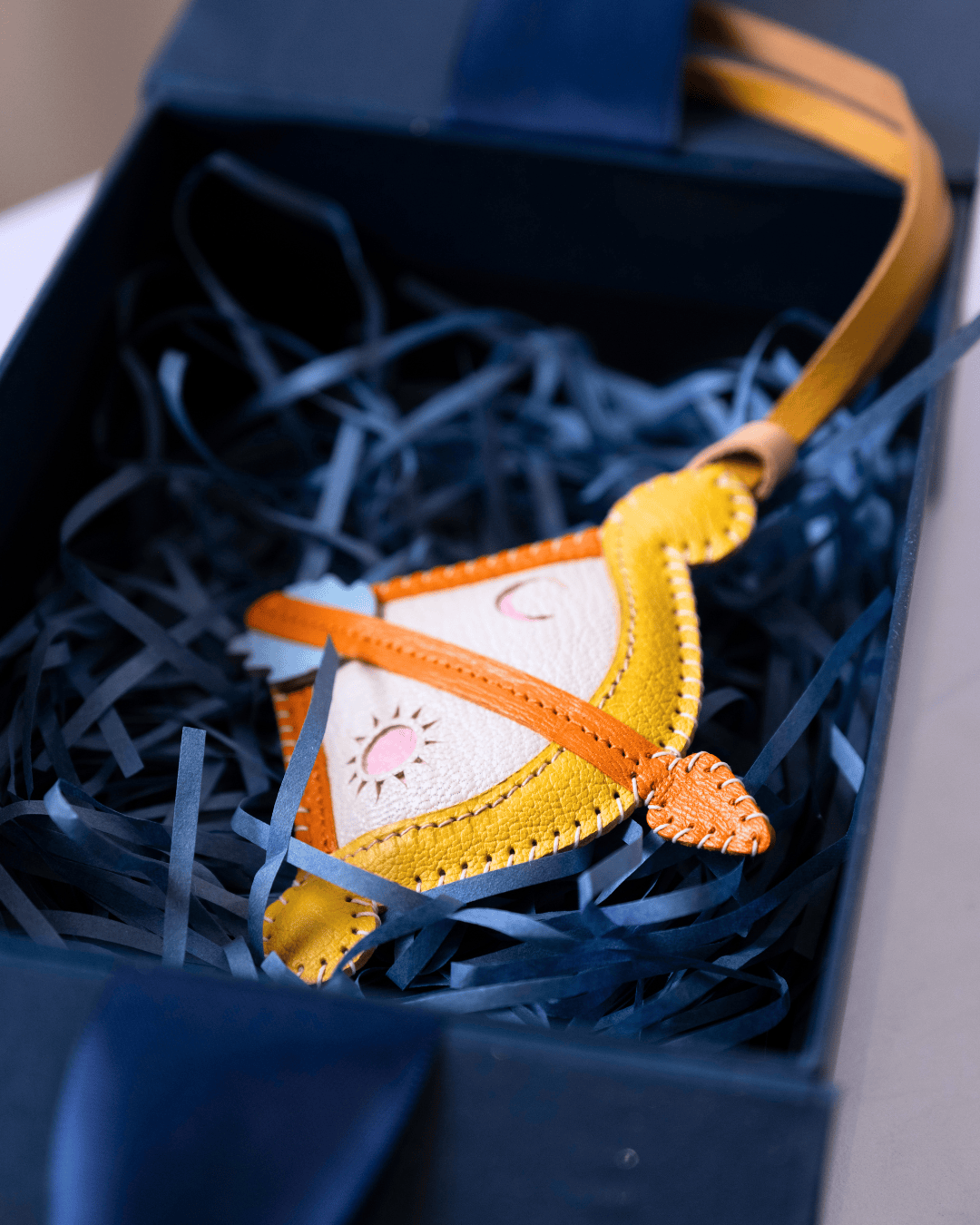 A small handmade felt ornament shaped like a smiling sun face, with orange trim, a cream center, and a yellow strap, resting in dark blue shredded paper inside an open gift box.