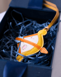 A small handmade felt ornament shaped like a smiling sun face, with orange trim, a cream center, and a yellow strap, resting in dark blue shredded paper inside an open gift box.
