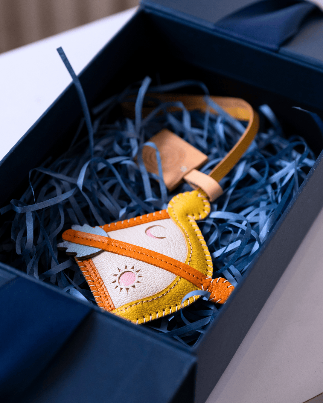 A small handmade leather pouch ornament with a yellow and cream body, orange trim, and a pink sun detail, nestled in blue shredded paper inside a dark navy gift box.