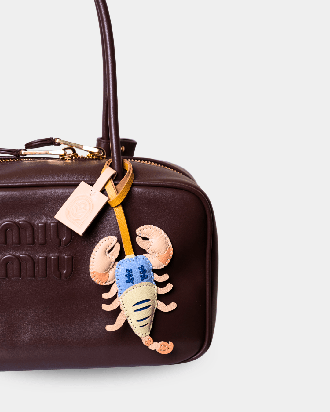 A close-up of a dark brown Miu Miu handbag featuring a decorative embroidered lobster charm in beige, blue, and yellow, with a gold-tone zipper and embossed logo.