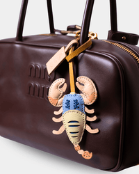 A close-up of a dark brown leather Miu Miu handbag featuring a decorative scorpion-shaped leather charm with a blue patterned body and tan claws, hanging from a gold-tone strap.