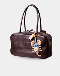 A dark brown leather Miu Miu handbag with a top handle, gold-tone zipper, and a dangling lobster charm, shown on a light gray background.