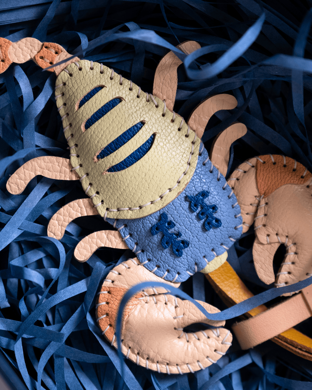 A handmade leather crab ornament or charm with tan and blue panels, photographed on blue paper shred.