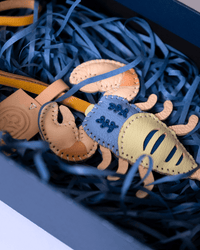 A close-up of a handmade leather crab ornament or charm inside a box filled with dark blue shredded paper. The crab features a light blue body with dark blue embroidered details, a pale yellow front section, and tan legs and claws.