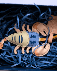 A handmade leather scorpion ornament or charm in natural tan and blue leather, nestled in blue shredded paper inside a dark box.