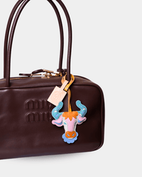 A close-up of a dark brown Miu Miu leather handbag featuring a colorful embroidered patch of a horned bull head with blue, pink, and orange details, and a small branded hangtag.