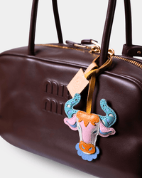 A close-up of a dark brown leather handbag with gold-tone hardware and a colorful embroidered bull head charm hanging from the zipper.