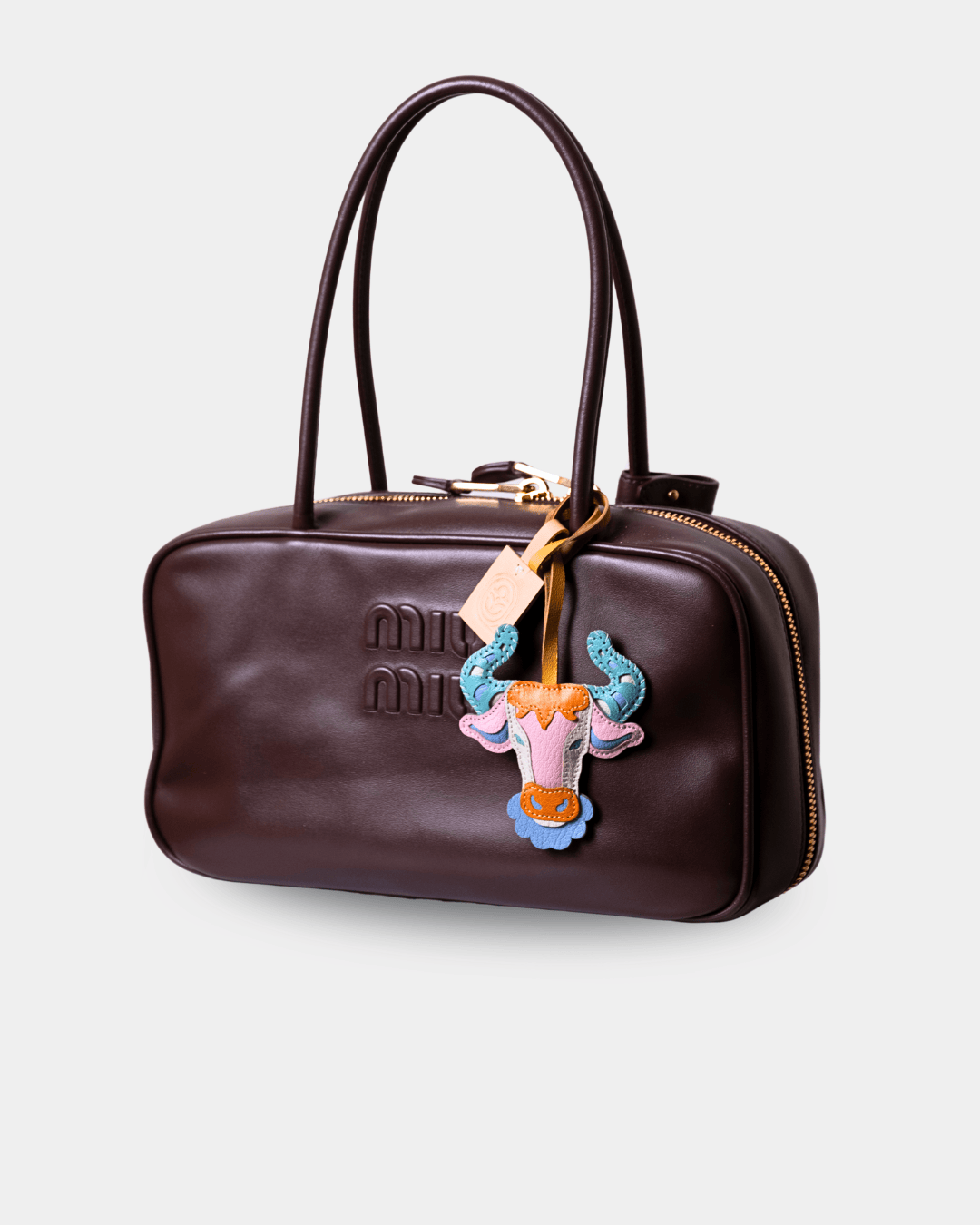 A dark brown leather handbag with double top handles, gold-tone hardware, and a colorful embroidered patch featuring a pink and blue horned animal design on the front.