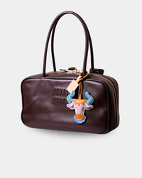 A dark brown leather handbag with double top handles, gold-tone hardware, and a colorful embroidered patch featuring a pink and blue horned animal design on the front.