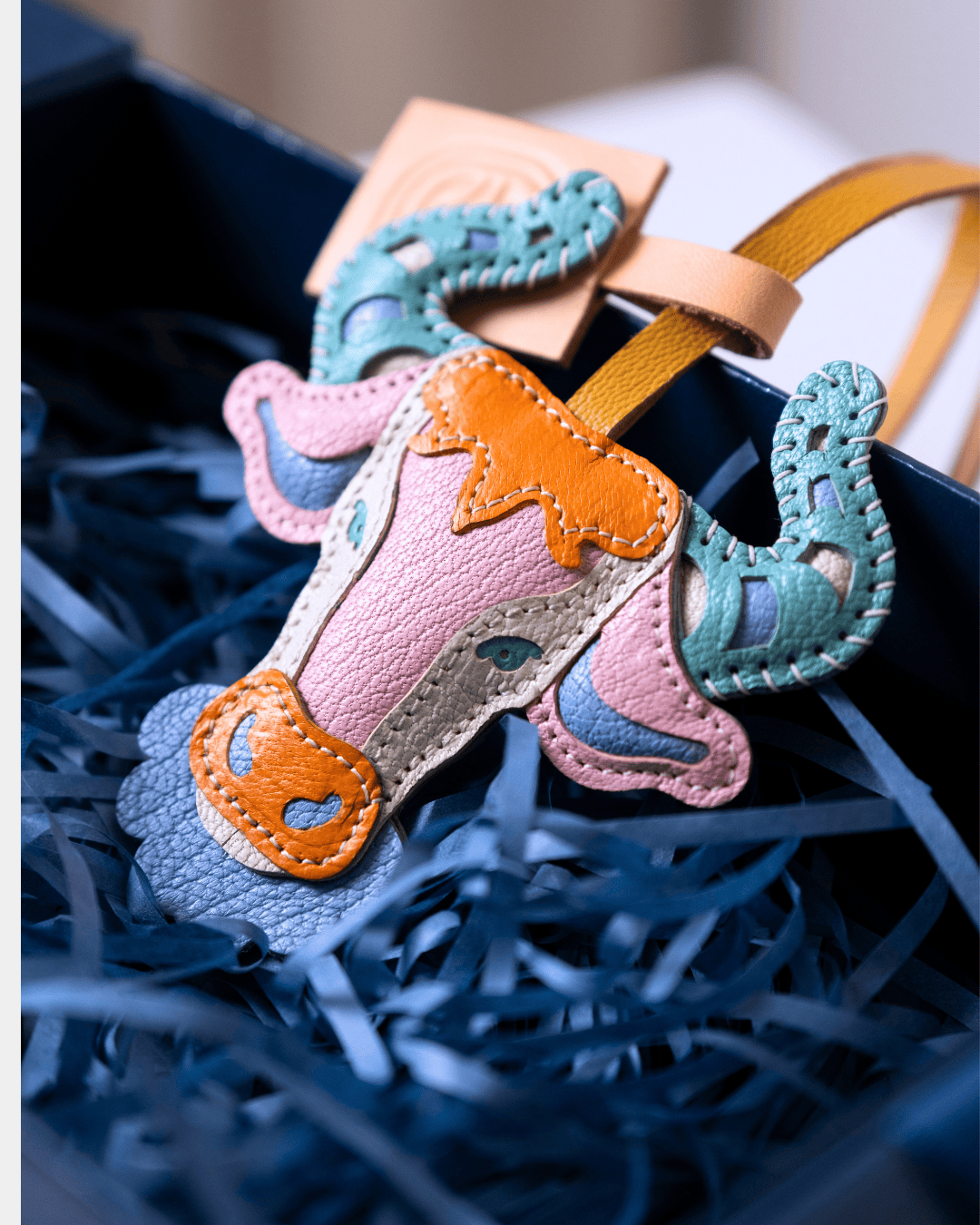 A colorful handmade leather cow-shaped bag charm or keychain resting in blue paper shred inside a dark box. The charm features a pink body, orange accents, and teal and blue decorative details.