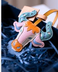 A colorful handmade leather cow-shaped bag charm or keychain resting in blue paper shred inside a dark box. The charm features a pink body, orange accents, and teal and blue decorative details.