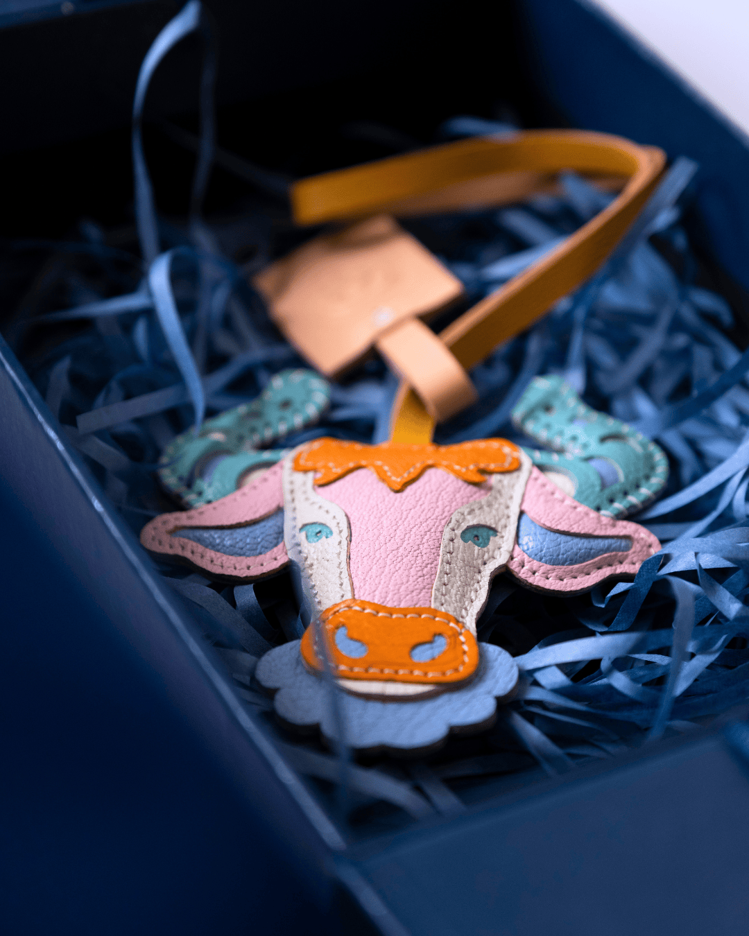 A handmade felt cow-shaped ornament with a yellow strap, resting in blue shredded paper inside a dark blue gift box.
