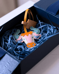 A colorful cow-shaped leather charm with a yellow strap, nestled in blue shredded paper inside a dark navy gift box.