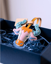 A handmade cow-shaped leather bag charm or keychain tag in pastel pink, orange, and teal, nestled in blue paper shred inside a dark navy gift box.