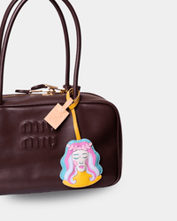 A close-up of a dark brown leather Miu Miu handbag featuring an embroidered patch of a woman's face with pink and blue hair, hanging from a yellow strap.