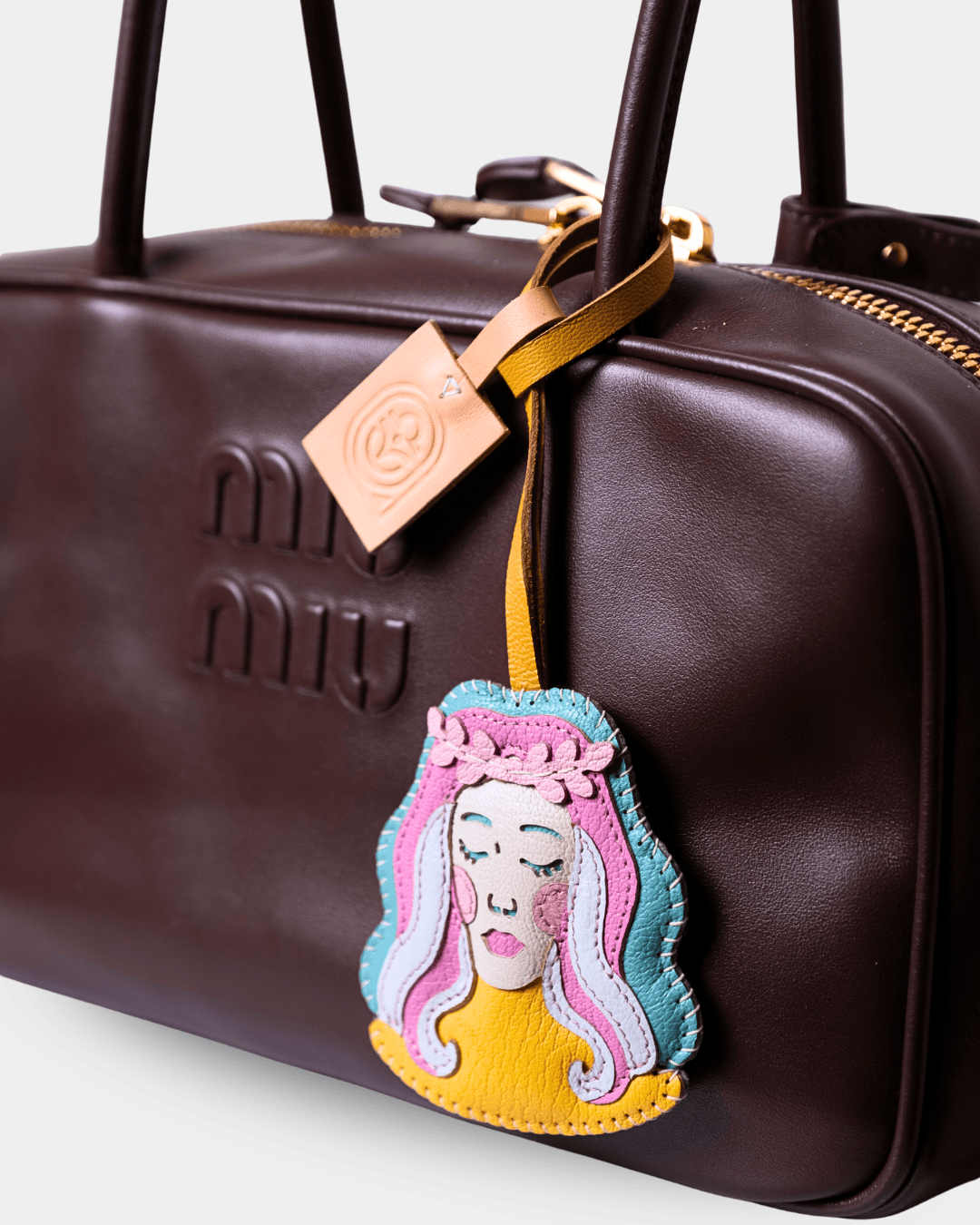 A close-up of a dark brown Miu Miu leather handbag featuring a colorful embroidered character charm with pink, blue, yellow, and white details, and a tan leather hangtag.
