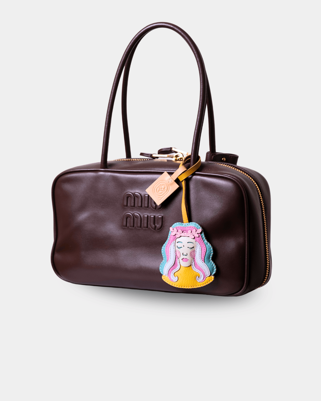 A dark brown faux-leather Miu Miu handbag with gold-tone hardware, featuring an embossed logo, dual top handles, and a colorful embroidered patch of a cartoon character hanging from the zipper.