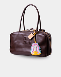 A dark brown faux-leather Miu Miu handbag with gold-tone hardware, featuring an embossed logo, dual top handles, and a colorful embroidered patch of a cartoon character hanging from the zipper.