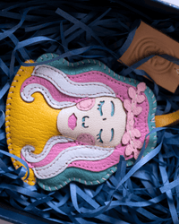A colorful handmade leather mermaid charm with yellow hair, pink and white waves, and a teal outline, nestled in blue paper shred inside a gift box.