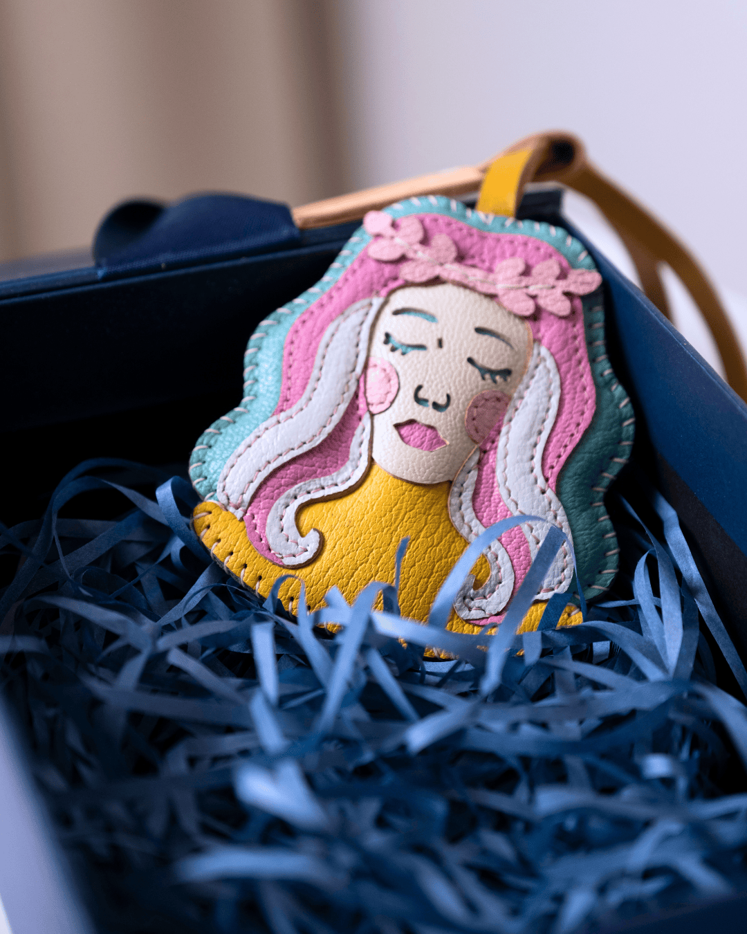 A colorful handmade leather patch featuring a stylized woman's face with long pink, blue, and white hair, resting in blue shredded paper inside a dark gift box.