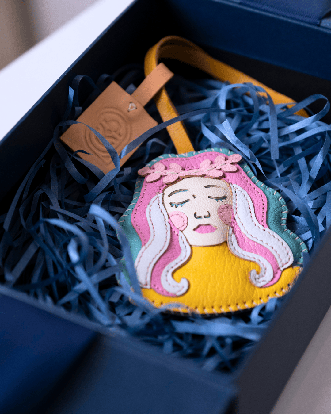 A handmade felt mermaid ornament with pink, white, yellow, and green colors, nestled in blue paper shred inside a dark blue gift box with a leather brand tag.